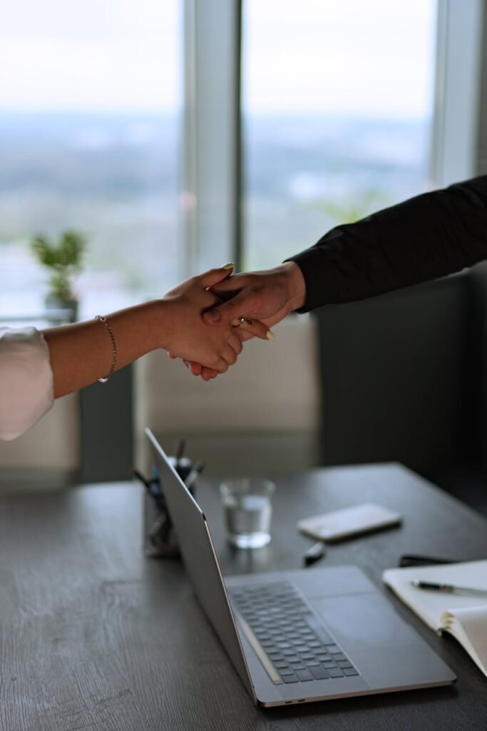 Business professionals shaking hands across a desk in a contemporary office with a laptop.